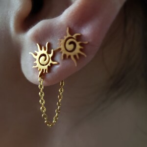 May include: Gold sun earrings with a chain connecting them. The earrings are in the shape of a sun with a spiral in the center.