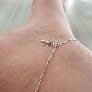 Puede incluir: Un tobillo de plata con un pequeño dije de dinosaurio. El tobillo está en el tobillo de una persona.