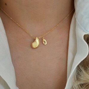 May include: A gold necklace with a nautilus shell charm and a small initial charm. The necklace is worn by a person with blonde hair.