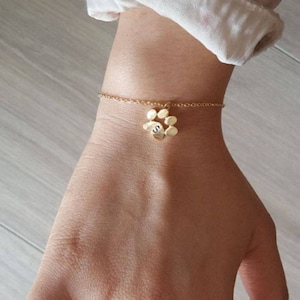 Puede incluir: Pulsera de oro con un dije en forma de huella de pata y una letra 'S' en el dije. La pulsera está en la muñeca.