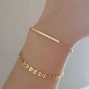 May include: Two gold bracelets on a wrist. One bracelet is a thin chain with a long, flat bar charm. The other bracelet is a chain with small, round, gold discs.