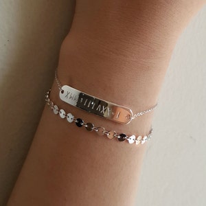 May include: Two silver bracelets on a person's wrist. The top bracelet has a rectangular tag with the Roman numeral 'MMXXI' engraved on it. The bottom bracelet is a chain with small silver circles.