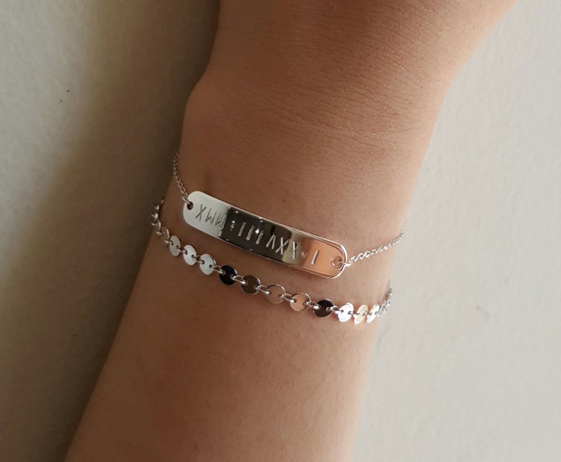 May include: Two silver bracelets on a person's wrist. The top bracelet has a rectangular tag with the Roman numeral 'MMXXI' engraved on it. The bottom bracelet is a chain with small silver circles.