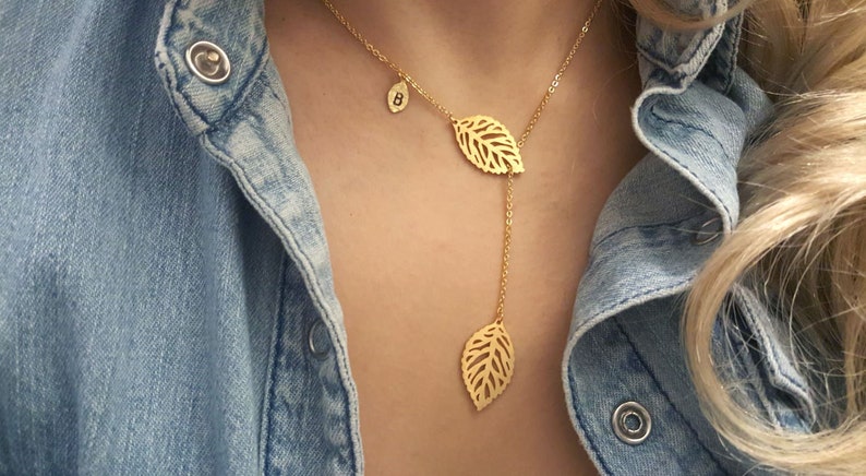 K&ouml;nnte beinhalten: Eine goldene Halskette mit einer zarten Kette und drei Blattanh&auml;ngern. Die Bl&auml;tter sind ausgeschnitten und haben ein filigranes Design. Die Halskette wird von einer Person mit einem Jeanshemd getragen.