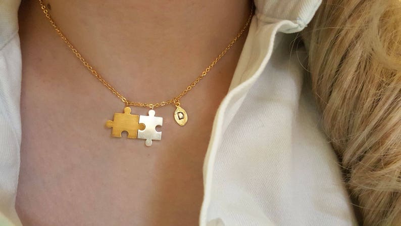 May include: A gold chain necklace with a two-piece puzzle pendant. One piece is gold and the other is silver. The pendant is attached to a small gold charm with the letter 'D' on it.