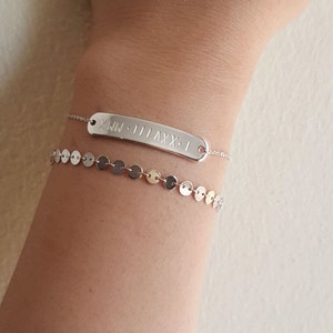 May include: A silver bracelet with a rectangular charm engraved with Roman numerals. The charm is attached to a delicate chain. The bracelet is layered with a second silver bracelet featuring a chain of small, round discs.