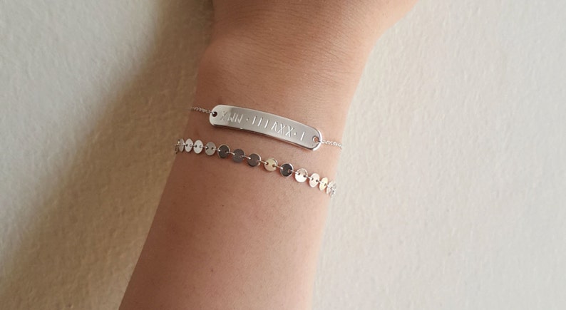 May include: A silver bracelet with a rectangular charm engraved with Roman numerals. The charm is attached to a delicate chain. The bracelet is layered with a second silver bracelet featuring a chain of small, round discs.