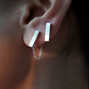 May include: Silver bar earrings with a delicate chain connecting them. The earrings are worn in the upper earlobe.
