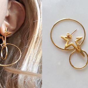 Puede incluir: Pendientes de aro dorados con un diseño de estrella. La estrella está unida a un aro más pequeño que cuelga del aro más grande.