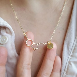 Op de afbeelding: Een gouden ketting met een hanger in de vorm van de chemische structuur van dopamine. De hanger heeft een kleine ronde bedel met de letter "R" erop gegraveerd.
