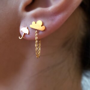 Könnte beinhalten: Goldfarbene Ohrringe mit einem Schirm- und Wolken-Design. Der Wolkenohrring hat eine Kette, die herunterhängt. Die Ohrringe werden am Ohr einer Person getragen.