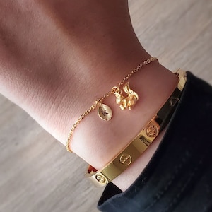 May include: A gold chain bracelet with a small leaf charm and a squirrel charm. The bracelet is worn on a wrist with a gold bangle bracelet.