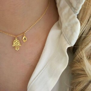 May include: A gold chain necklace with a hamsa hand charm and a small initial charm. The initial charm is engraved with the letter "A".