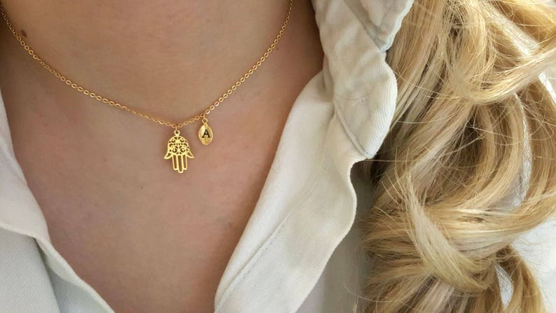 May include: A gold chain necklace with a hamsa hand charm and a small initial charm. The initial charm is engraved with the letter "A".