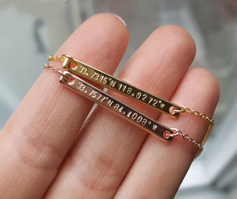 May include: Two gold bar necklaces with coordinates engraved on them. The top necklace has the coordinates "33. 7315' N 118. 0272' W" and the bottom necklace has the coordinates "33. 1517' N 84. 4008' W".