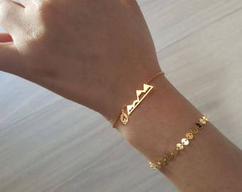 Könnte beinhalten: Zwei goldene Armbänder am Handgelenk einer Person. Ein Armband hat einen Anhänger in Form einer Bergkette, das andere ist eine Kette aus kleinen goldenen Kreisen.