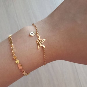 May include: Two gold bracelets are shown on a wrist. One is a delicate chain with small, round charms. The other features a geometric bird charm and a small initial charm. The bracelets are made of a shiny, gold-colored metal.