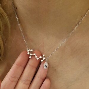May include: A silver necklace with a constellation pendant and a small charm with the letter A. The pendant is made of small silver squares with clear crystals.