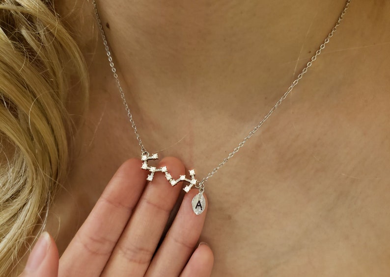 May include: A silver necklace with a constellation pendant and a small charm with the letter A. The pendant is made of small silver squares with clear crystals.