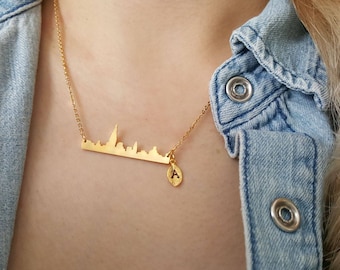 Puede incluir: Un collar de oro con un colgante de ciudad y un pequeño encanto inicial. El collar lo lleva una persona que lleva una camisa vaquera.