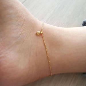 K&ouml;nnte beinhalten: Ein goldenes Fu&szlig;kettchen mit einem kleinen Ananas-Anh&auml;nger. Das Fu&szlig;kettchen ist an einem Fu&szlig;gelenk.