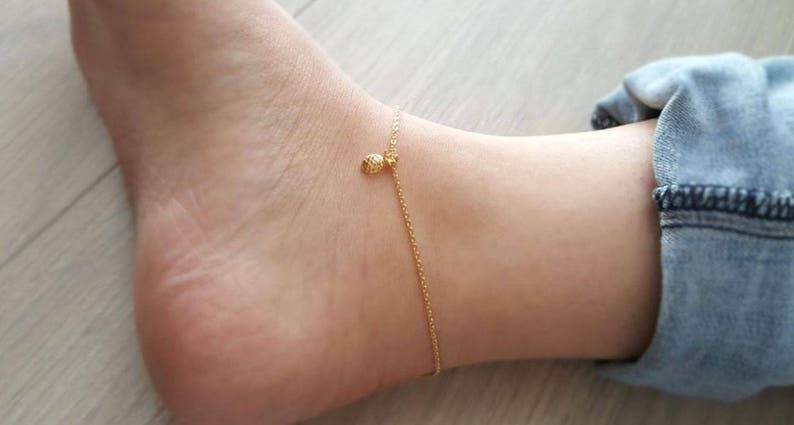 K&ouml;nnte beinhalten: Ein goldenes Fu&szlig;kettchen mit einem kleinen Ananas-Anh&auml;nger. Das Fu&szlig;kettchen ist an einem Fu&szlig;gelenk.