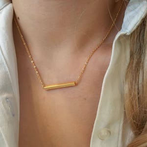 May include: A gold bar necklace with a delicate chain. The necklace is worn by a person with long blonde hair.
