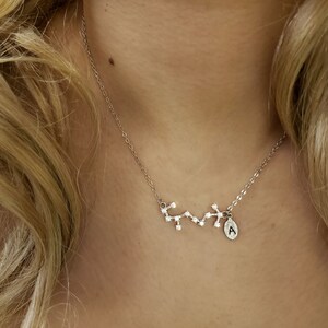 May include: A silver necklace with a constellation charm and a small letter 'A' charm. The necklace is on a person's neck.