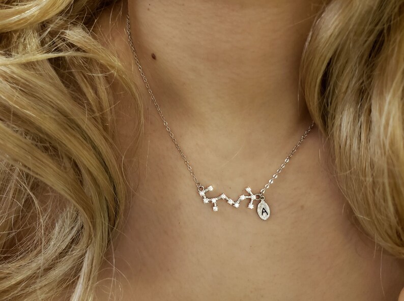 May include: A silver necklace with a constellation charm and a small letter 'A' charm. The necklace is on a person's neck.