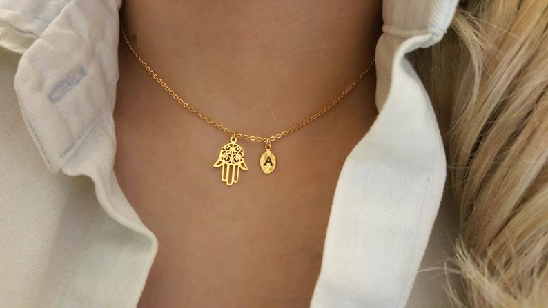 May include: A gold chain necklace with two charms: a hamsa hand and a letter 'A'.