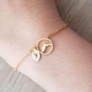 May include: A gold chain bracelet with a small leaf charm and a larger charm shaped like a mountain range inside a circle.