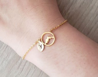 Könnte beinhalten: Ein goldenes Kettenarmband mit einem kleinen Blatt-Anhänger und einem größeren Anhänger in Form einer Bergkette in einem Kreis.