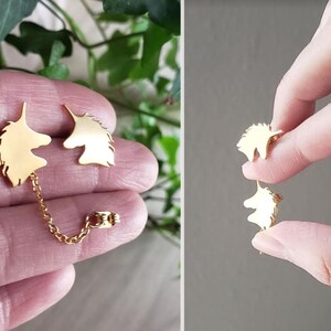 May include: Gold unicorn silhouette earrings with a delicate chain connecting the two pieces. The earrings are shown on a hand.