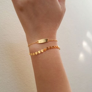 May include: Two gold bracelets on a wrist. The top bracelet has a small rectangular tag with the letters "J" and "S" and a heart symbol engraved on it. The bottom bracelet is a chain of small gold circles.