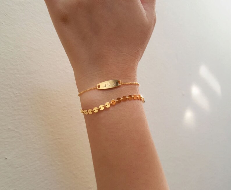 May include: Two gold bracelets on a wrist. The top bracelet has a small rectangular tag with the letters "J" and "S" and a heart symbol engraved on it. The bottom bracelet is a chain of small gold circles.