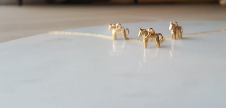 May include: Three gold-colored Dala horse charms on a delicate chain necklace. The charms are detailed with traditional Swedish Dala horse designs.
