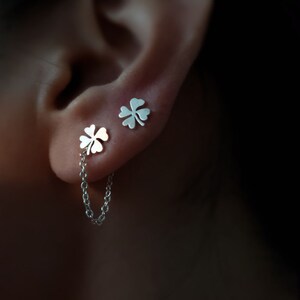 以下が含まれることがあります： 繊細なチェーンで繋がった、銀製の四つ葉のクローバーのイヤリングのペア。イヤリングは耳の上の軟骨につけられています。