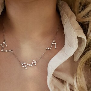May include: A silver necklace with three constellation pendants. The pendants are in the shape of Sagittarius, Libra, and Aquarius. The necklace is on a model wearing a white shirt.