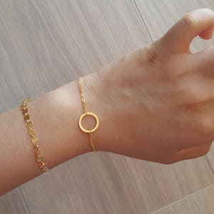 May include: Two gold bracelets on a person's wrist. One bracelet is a delicate chain with a circle pendant. The other bracelet is a chain of small gold discs.