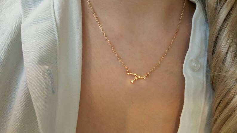 May include: A gold chain necklace with a delicate constellation pendant. The pendant is shaped like the constellation Gemini.