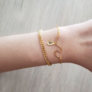 Può includere: Due braccialetti d'oro su un polso. Un braccialetto è una semplice catena, l'altro ha un ciondolo con la lettera 'S' in corsivo.