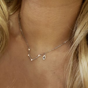 May include: A silver necklace with a small pendant featuring the letter "A" and a constellation design. The necklace is worn by a person with long blonde hair.