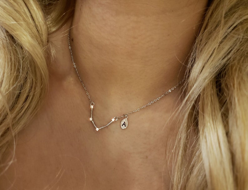 May include: A silver necklace with a small pendant featuring the letter "A" and a constellation design. The necklace is worn by a person with long blonde hair.