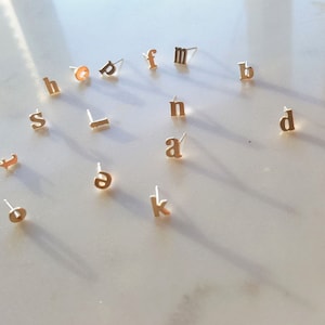 Puede incluir: Una colección de pendientes de letras doradas, incluyendo las letras a, b, d, f, h, k, l, m, n, s y t. Los pendientes están dispuestos sobre una superficie blanca.
