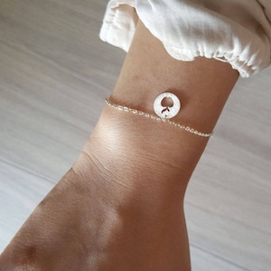 May include: A delicate silver bracelet with a round charm featuring a spade design. The bracelet is worn on a wrist, showcasing the jewelry's fine details and elegant simplicity. The bracelet is a stylish accessory.
