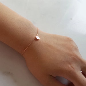 Può includere: Un delicato braccialetto in oro rosa con un piccolo ciondolo quadrato con un'iniziale. Il braccialetto è indossato su un polso, mettendo in risalto la sua catena sottile e il design minimalista. Il ciondolo è un punto focale, aggiungendo un tocco personalizzato al gioiello.