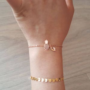 May include: A gold bracelet with a pineapple charm and a small leaf charm. The bracelet is on a person's wrist.