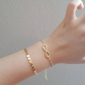 Puede incluir: Dos pulseras doradas se muestran en una muñeca. Una presenta una serie de pequeños discos redondos, mientras que la otra muestra un símbolo de infinito. Las pulseras son delicadas y parecen estar hechas de una cadena fina.