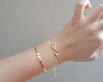 Peut inclure: Deux bracelets dorés sont présentés sur un poignet. L'un est orné de petits disques ronds, l'autre d'un symbole infini. Les bracelets sont délicats et semblent être faits d'une fine chaîne.