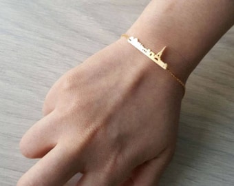 Könnte beinhalten: Ein goldfarbenes Armband mit einer zarten Kette und einem Anhänger mit der Skyline von Paris, einschließlich des Eiffelturms.